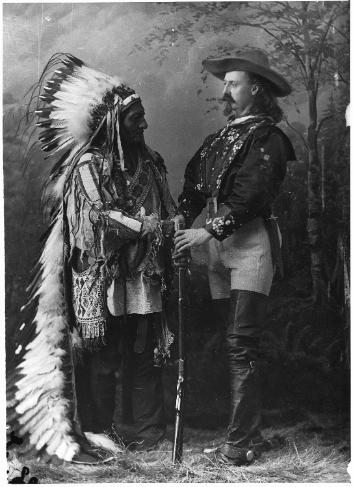 Photo en noir et blanc de deux hommes debout, l'un en tenue d'apparat des Indiens Lakotas, l'autre en tenue de cowboy.